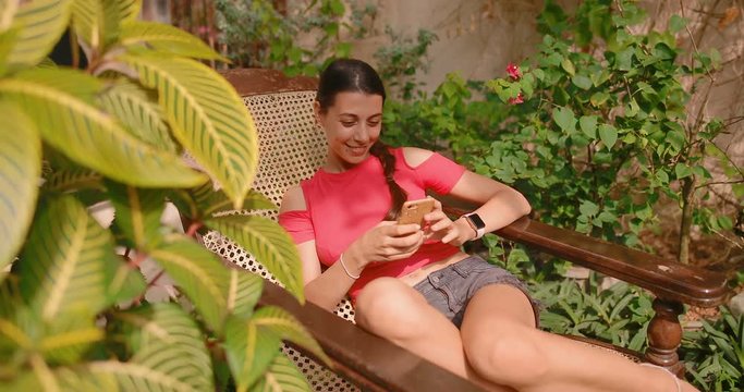 Attractive girl sitting in a deckchair in the garden uses a smartphone. The girl smiles and looks happy. tracking shot. in slow motion