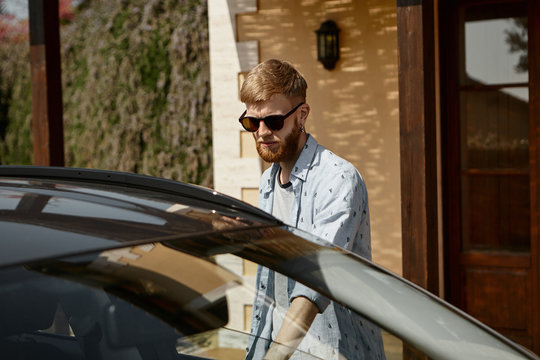 Attractive Bearded Hipster Guy In Black Shades Standing By Car With Arm Extended Going To Open Front Door And Seat In Driver's Seat. Unshaven Man Commuting To Work On Automobile In The Morning