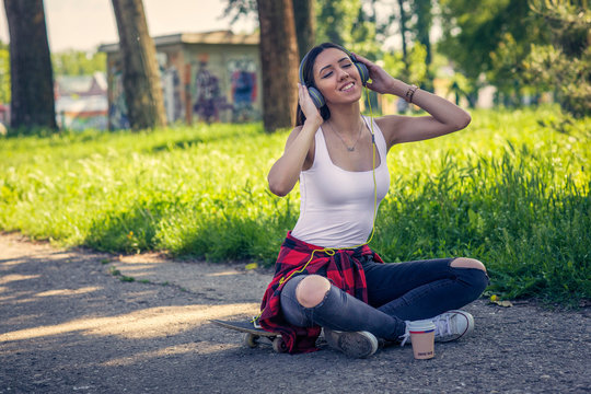 Smiling urban girl sitting on skateboard and listening music. Outdoors, urban lifestyle..