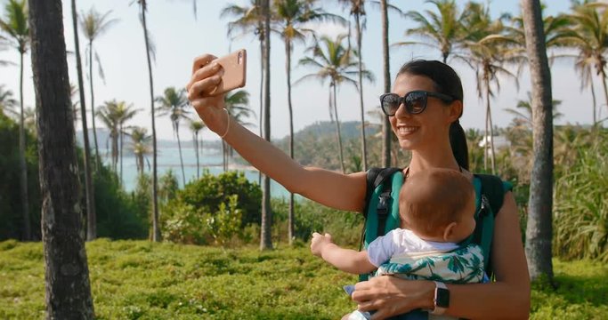 Modern mother with a baby in her arms is taking pictures of herself against the backdrop of palm trees and the ocean
