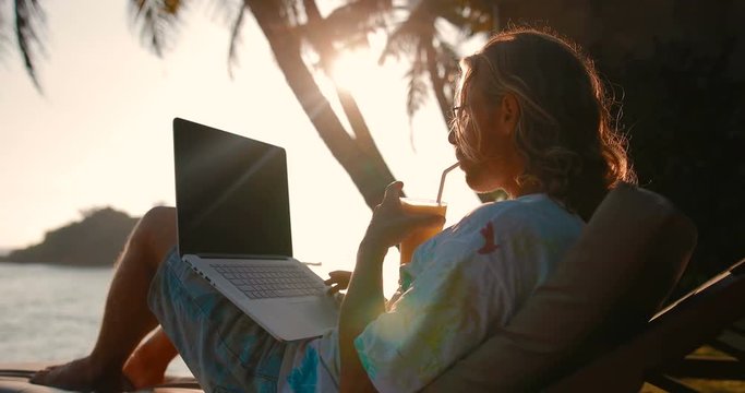 A young relaxed man working on the beach using a smartphone and laptop. man drinking fruit juice. tracking shot. in slow motion
