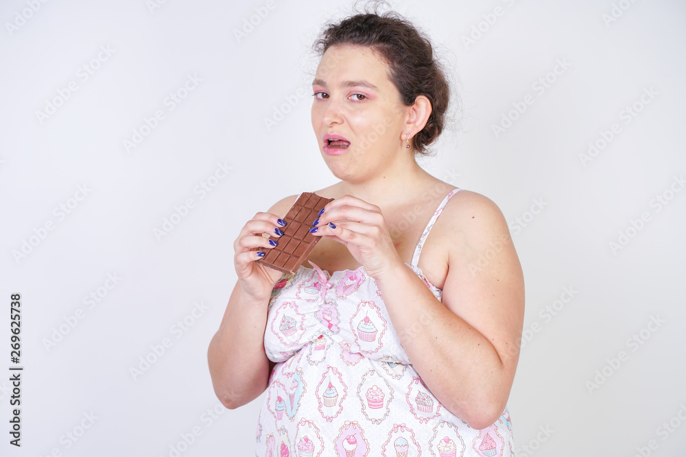 Wall mural curvy woman in funny pajamas with a chocolate bar stands on a white background in the Studio