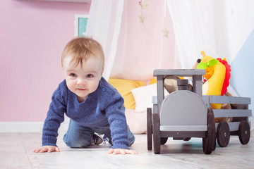 Boy blond in a blue sweater crawls on a wooden floor. One year old baby playing with wooden toys.