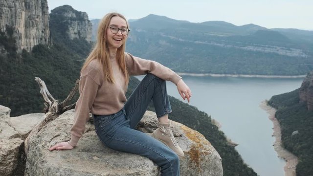 Happy attractive caucasian girl in glasses with a backpack behind her back laughs, sits on the edge of a cliff and enjoys nature