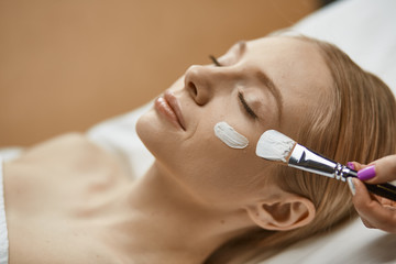 people, beauty, cosmetology and treatment concept - close up of beautiful young woman lying with closed eyes and cosmetologist applying facial mask by brush at spa