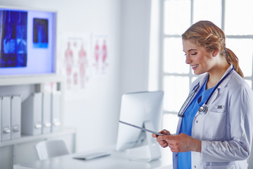 Young woman doctor is standing with board with clipboard smiling in hospital office