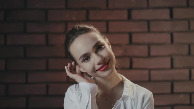 Stylish girl touching hair and ear while posing in studio with brick wall
