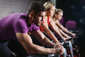 Close up hands of man biking in spinning class. Group of smiling friends at gym exercising on stationary bike. Happy cheerful athletes training on exercise bike. - obrazy, fototapety, plakaty