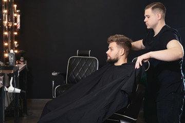Professonal barber ready to work and covering with cape bearded man on barbershop background....
