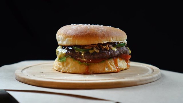 Fresh juicy burger with cheese, sauce and vegetables rotating on the wooden board on a black background