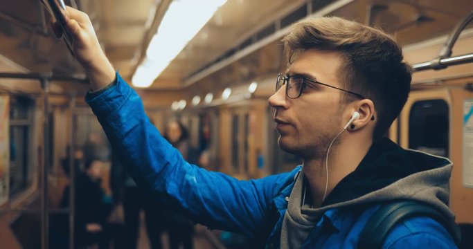 Passenger rides in the car and holds on to the rail, listens to music with headphones. Young man rides the subway, touches his hair with his hand.