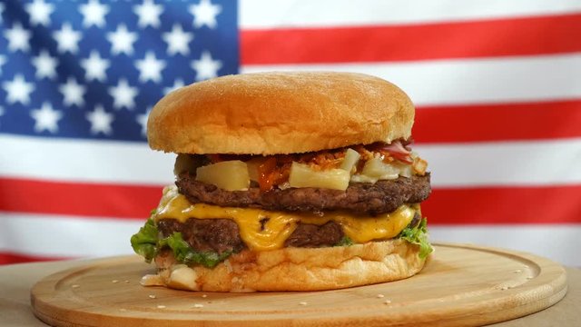 Fresh juicy burger with cheese, sauce and vegetables rotating on a wooden board on us flag background.