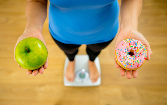 Woman On Scale Measuring Weight Holding Apple And Donuts Choosing Between Healthy Or Unhealthy Food