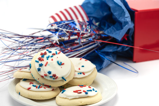 Red White And Blue Patriotic Frosted Sugar Cookies On A White Plate With Flag And Mylar Sparkly Decor Out Of Focus In The Background