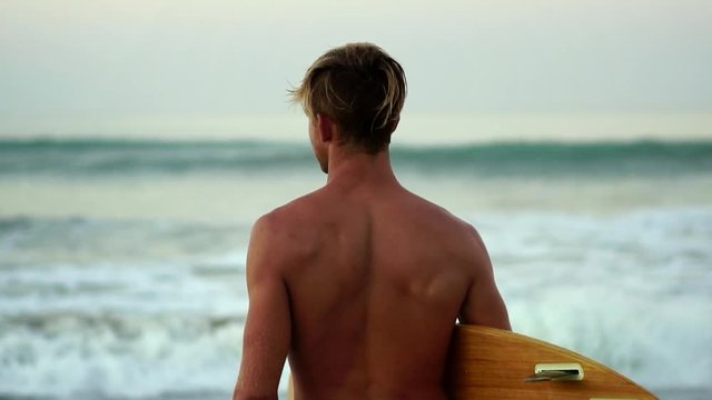 Beautiful blonde surfer man on beach holding surfboard. slow motion. Modern Healthy Active Lifestyle. Surfing. Summer Vacation