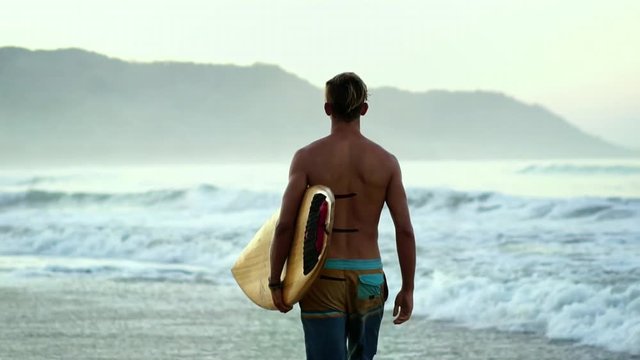 Young attractive surfer guy at beach with surfboard, summer surf adventure, free spirit lifestyle. Modern active sport & travel vacation. Slow Motion. 