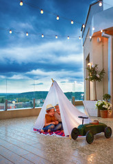happy kids are hiding in handmade wigwam from a sudden summer rain at the patio, playing together © Olesia Bilkei