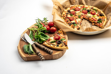 on a white background an isolated piece of pizza, on a wooden board, next to fresh basil leaves and tomatoes, in the back lies a frying pan with pizza in baking paper