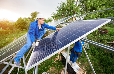Installing of stand-alone solar photo voltaic panel system. Two technicians in hard-hats mounting big shiny solar module on platform on green summer view background. Alternative energy concept.