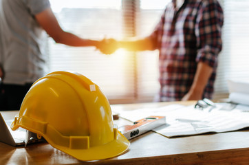 Fototapeta yellow safety helmet on workplace desk with construction worker team hands shaking greeting start up plan new project contract in office center at construction site, partnership and contractor concept obraz