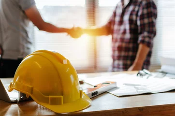Foto op Plexiglas yellow safety helmet on workplace desk with construction worker team hands shaking greeting start up plan new project contract in office center at construction site, partnership and contractor concept © Vittaya_25