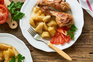 Baked chicken legs with potatoes and tomatoes in a white plate on a wooden background
