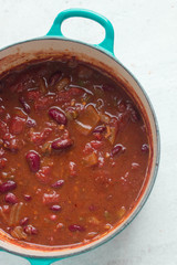 Top view of turquoise Le Creuset Dutch oven with chili sin carne made of kidney beans, tomatoes, onions, garlic served with a spoon and cilantro on light natural stone background. Copy space, close up