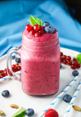 Healthy frozen strawberry and raspberry smoothie in a mason jar glass with a straw, fresh mint and scattered berries: currants, blueberries and blue straws on a light white plate with a blue napkin. 