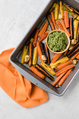 Top view and close up of a baking/frying sheet with roasted/baked rainbow orange, yellow and purple carrots with garlic served with basil, parmesan and olive oil green pesto with an orange napkin. 