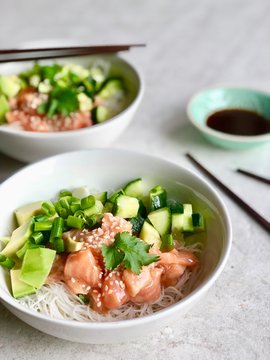 Side view of two Poke bowls with raw marinated salmon, cilantro, avocado, cucumber , sesame seeds and rice noodles served with soy/tamari sauce and wooden chopsticks. Flat lay on light background. 