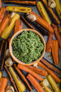 Top view and close up of a baking/frying sheet with roasted/baked rainbow orange, yellow and purple carrots with garlic served with basil, parmesan and olive oil green pesto with an orange napkin. 