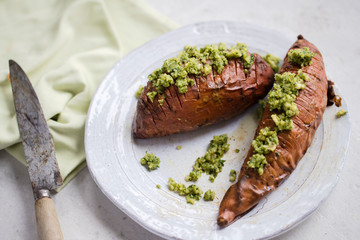 Top view of two hasselback baked sweet potatoes served with green walnut, kale and parsley pesto on white plate with rustic knife and light green napkin. Top view, flat lay, close up. 