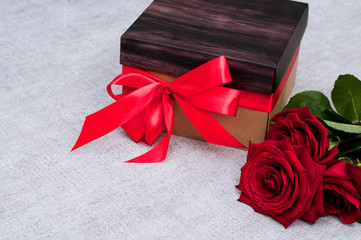 A beautiful gift box with a red ribbon along with fresh red roses and empty space for your design or montage. The concept of holiday and gifts.