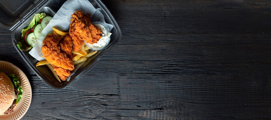 Hamburger, french fries and fried chicken in takeaway containers on the wooden background. Food delivery and fast food concept. Banner