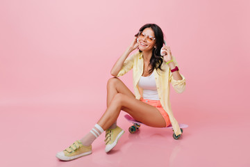 Refined hispanic girl wears yellow sneakers and jacket holding big headphones and laughing. Indoor...