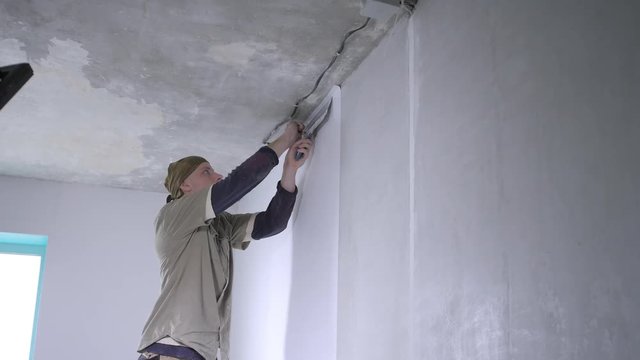skilled handyman cuts excess wallpaper pieces near ceiling with metal spatula and box cutter