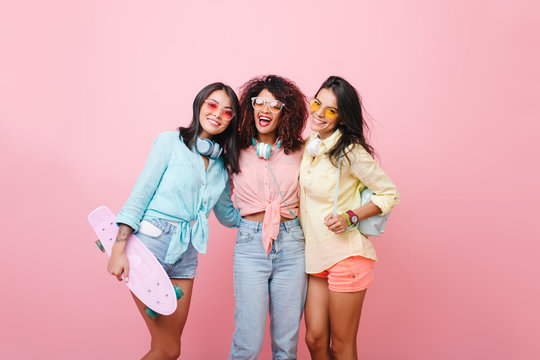 Graceful girl in yellow shirt with leather backpack posing near african curly friend in jeans. Cheerful black young woman in sunglasses standing between european and latino ladies on pink background.