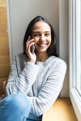 I am waiting for you. Beautiful young woman talking on mobile phone with smile while sitting at windowsill