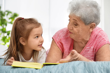 Cute girl and her grandmother reading book on bed at home