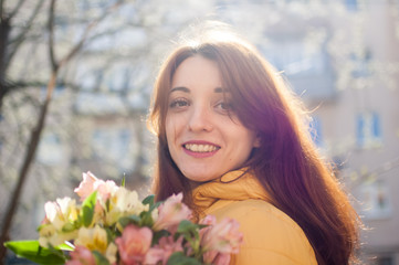 Fototapeta premium Outdoors female portrait of attractive brunette girl in yellow jacket holding a big bouquet of colorful flowers enjoying spring and looking at tne camera on building background