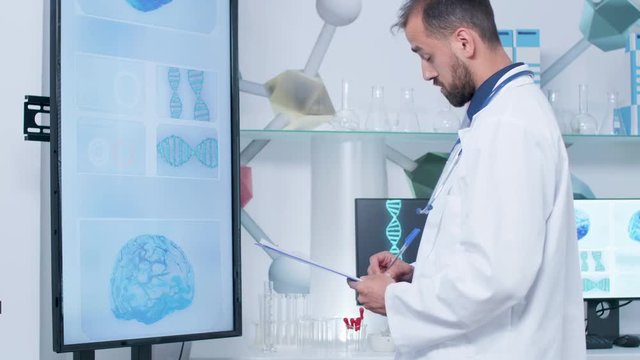 Doctor in his modern office or research center looking at big screen TV with 3D brain models and animated DNA strings. He is taking notes in the clipboard