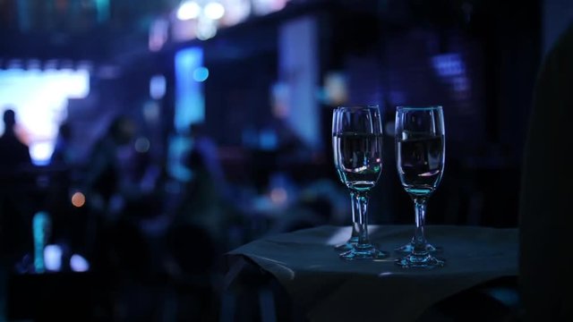 waiter holds tray with white napkin and glass goblets in crowded night club with coloured light close view