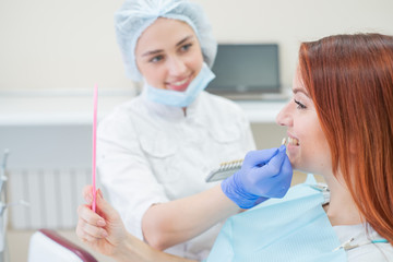 Obraz na płótnie Canvas Check and select the color of the teeth in the dentists chair. Dentist makes the process of treating a beautiful young redhaired girl. Selection of a dental implant. Patient looking in the mirror.