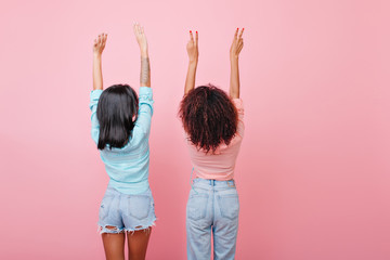 Indoor photo from back of stretching brunette girls in jeans. Glamorous dark-haired ladies in vintage attire posing with hands up on pink background.