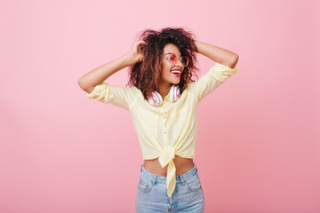 Slim young woman with brown skin looking away with sincere smile on light background. Sensual woman plays with soft curly hair and expressing happy emotions.