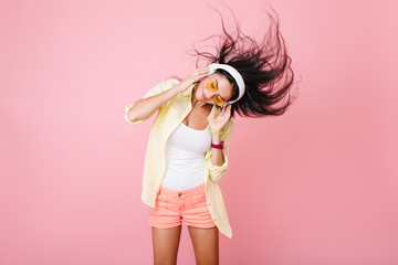 Joyful tanned woman in pink bracelet listening music and waving head. Indoor portrait of beautiful asian female model in orange sunglasses chilling during studio photoshoot.