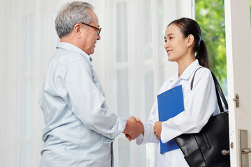Obraz premium Asian female doctor with blue medical card talking to her senior patient and shaking his hand while they standing near entrance of the house