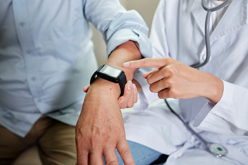 Obraz na płótnie Canvas Close-up of female doctor taking pulse of senior patient she pointing at wristwatch and checking the time