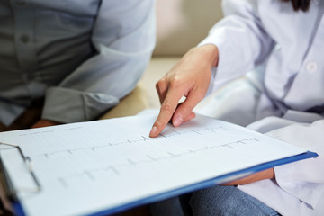 Close-up of cardiologist pointing at clipboard with cardiogram and showing it to senior patient during his visit at hospital