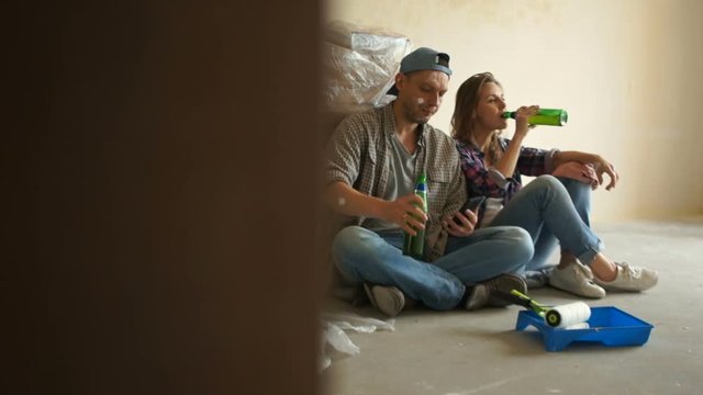 A man and a woman took a break during home renovation. Newlyweds drink beer from bottles while sitting on the floor of their new apartment. Rest after repair